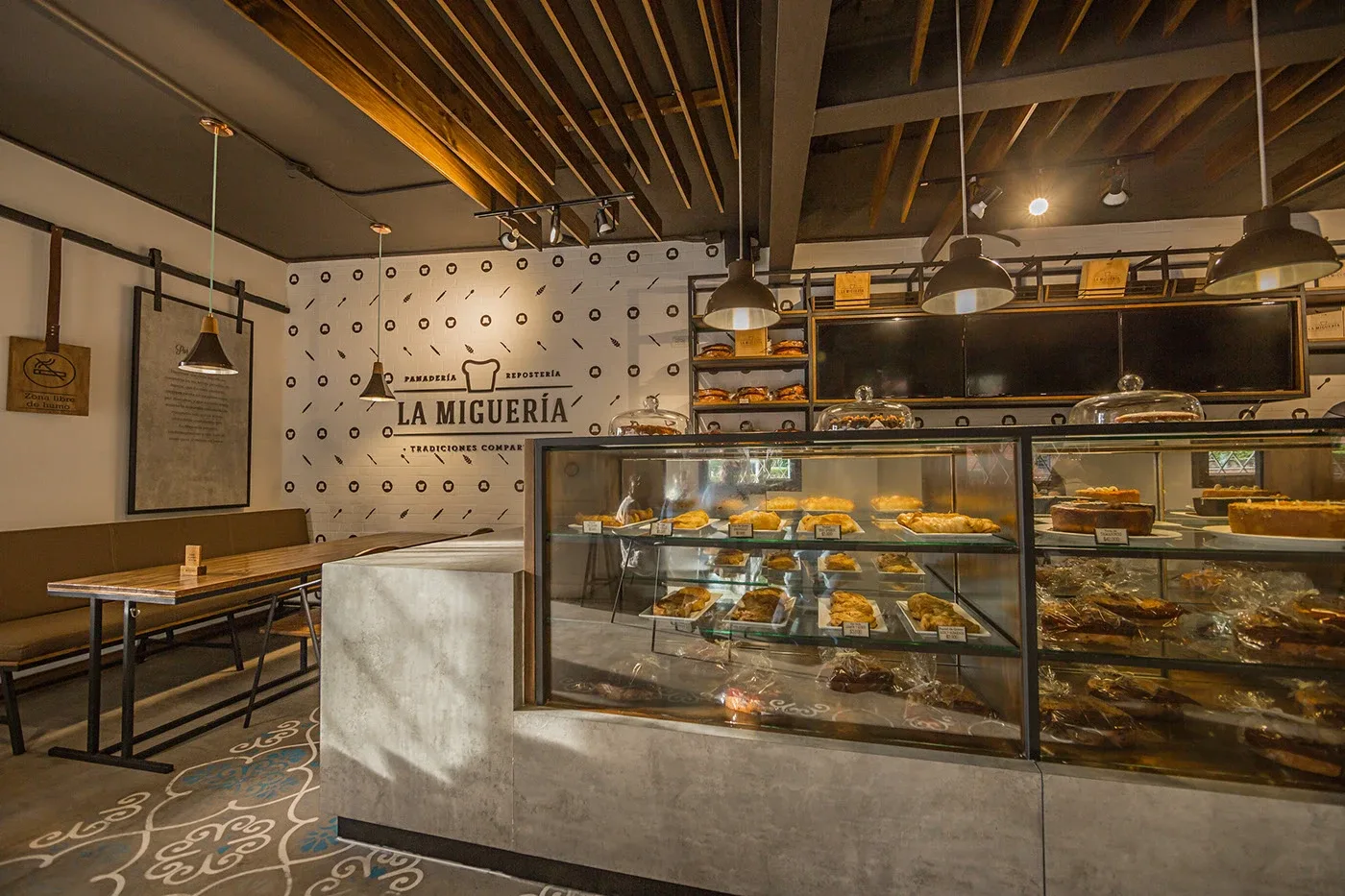 Interior of La Miguería bakery with a glass display case full of pastries and rustic wooden industrial décor.