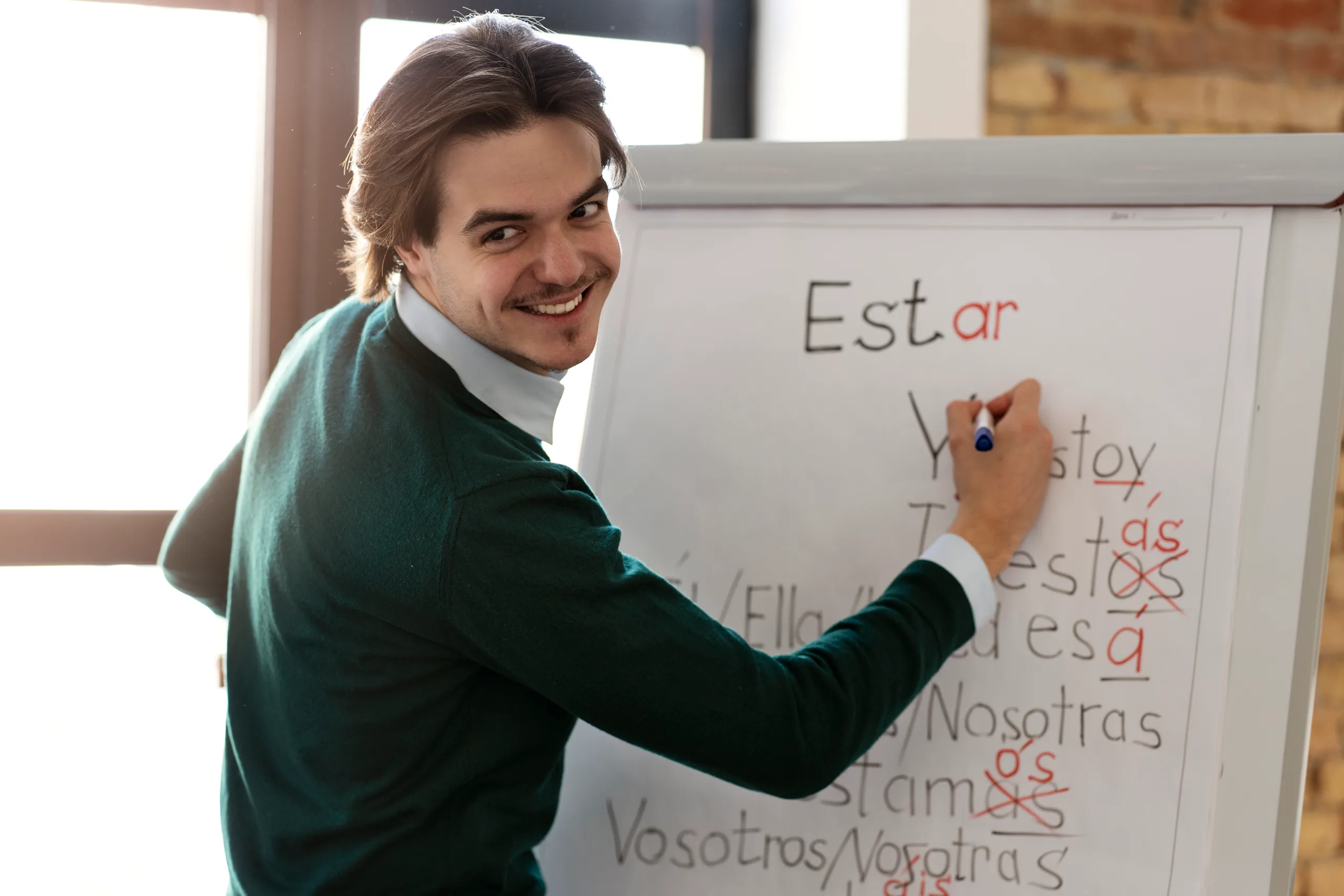 A teacher in a green sweater writes the conjugation of the Spanish verb estar on a whiteboard.