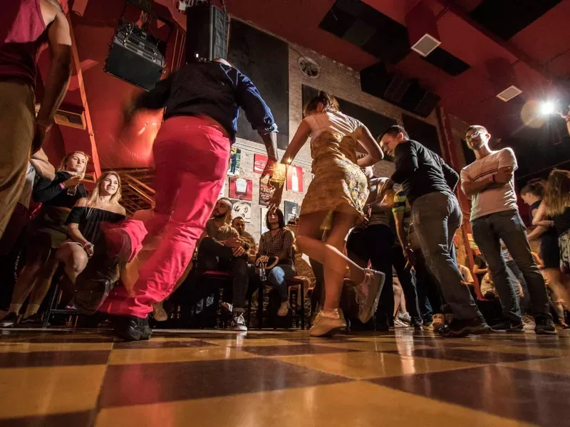 People dancing energetically in a warmly lit red-walled dance hall with a checkered floor.