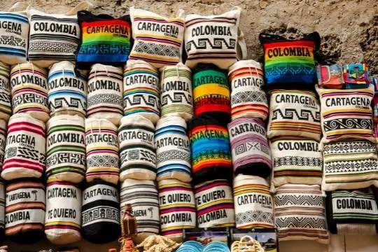 Handmade cushions and bags stacked at a market stall, with colorful geometric patterns and the names of Colombian cities embroidered on them.