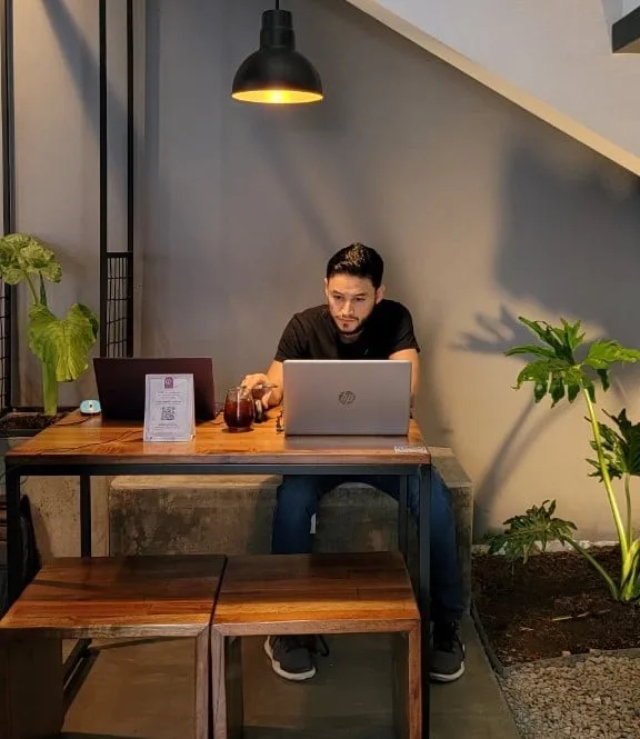 Un hombre joven con camiseta negra trabaja concentrado frente a un portátil HP plateado sobre una mesa de madera con estructura metálica en un café de estilo industrial, iluminado