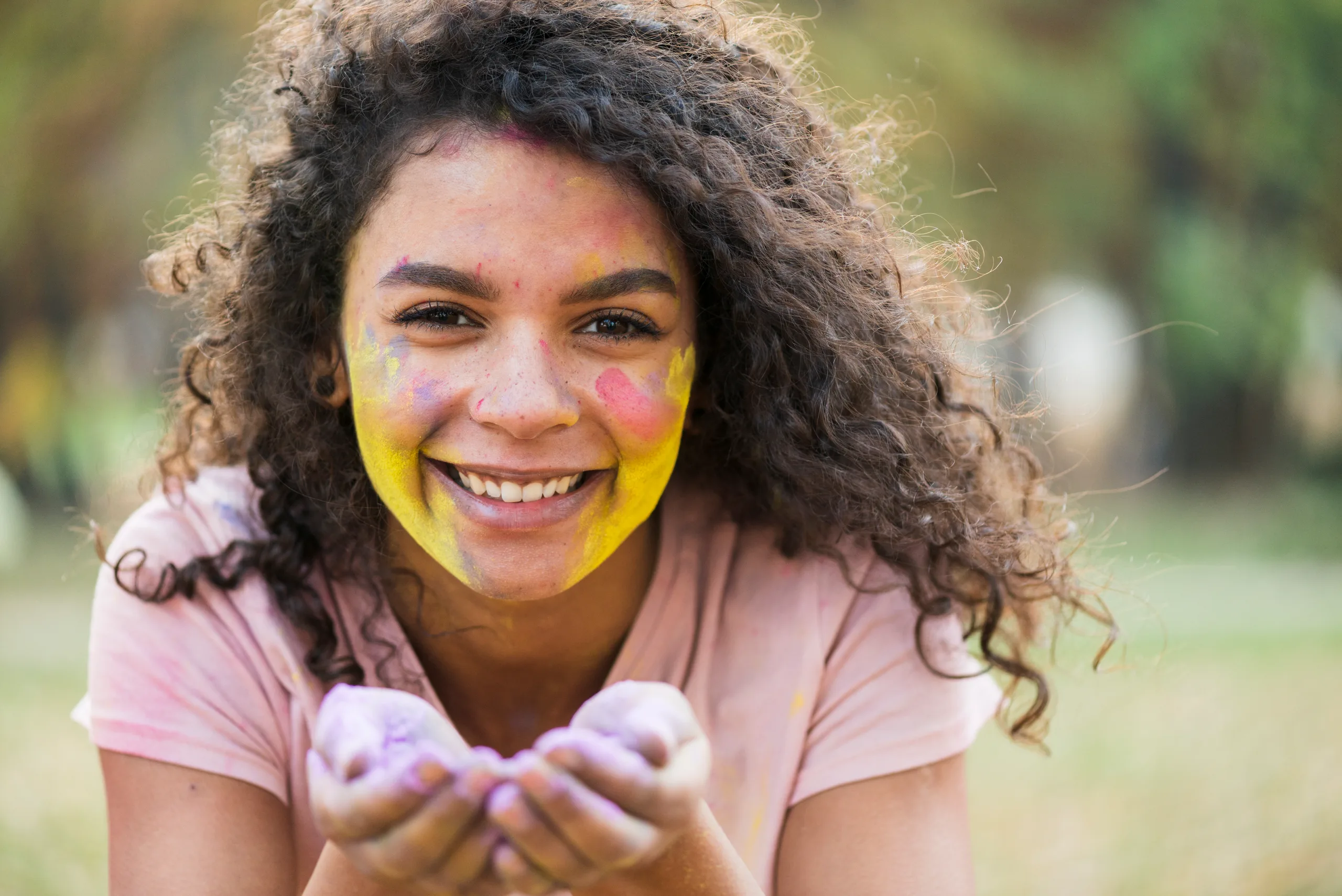 A smiling young woman with curly hair, her face covered in colored powder, holding more lilac powder in her cupped hands.