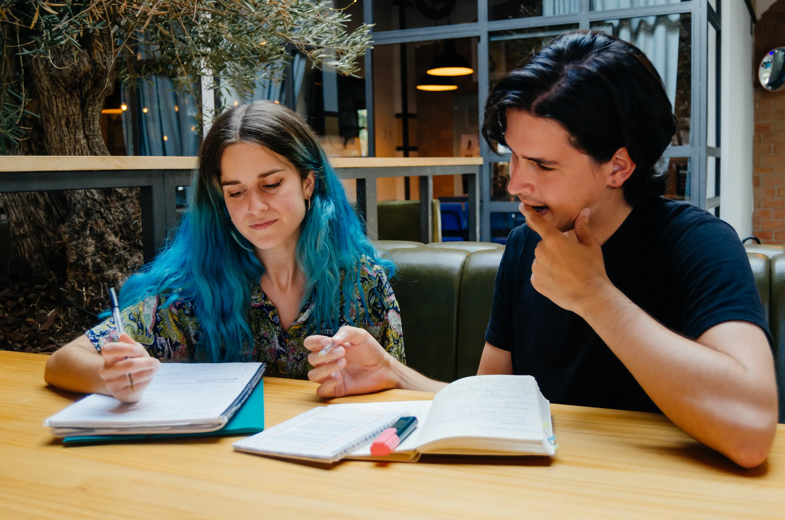 Dos jóvenes estudiantes —una chica con el pelo azul y un chico con camiseta negra— sentados juntos en una mesa de madera clara, estudiando: ella escribe en un cuaderno y él consult