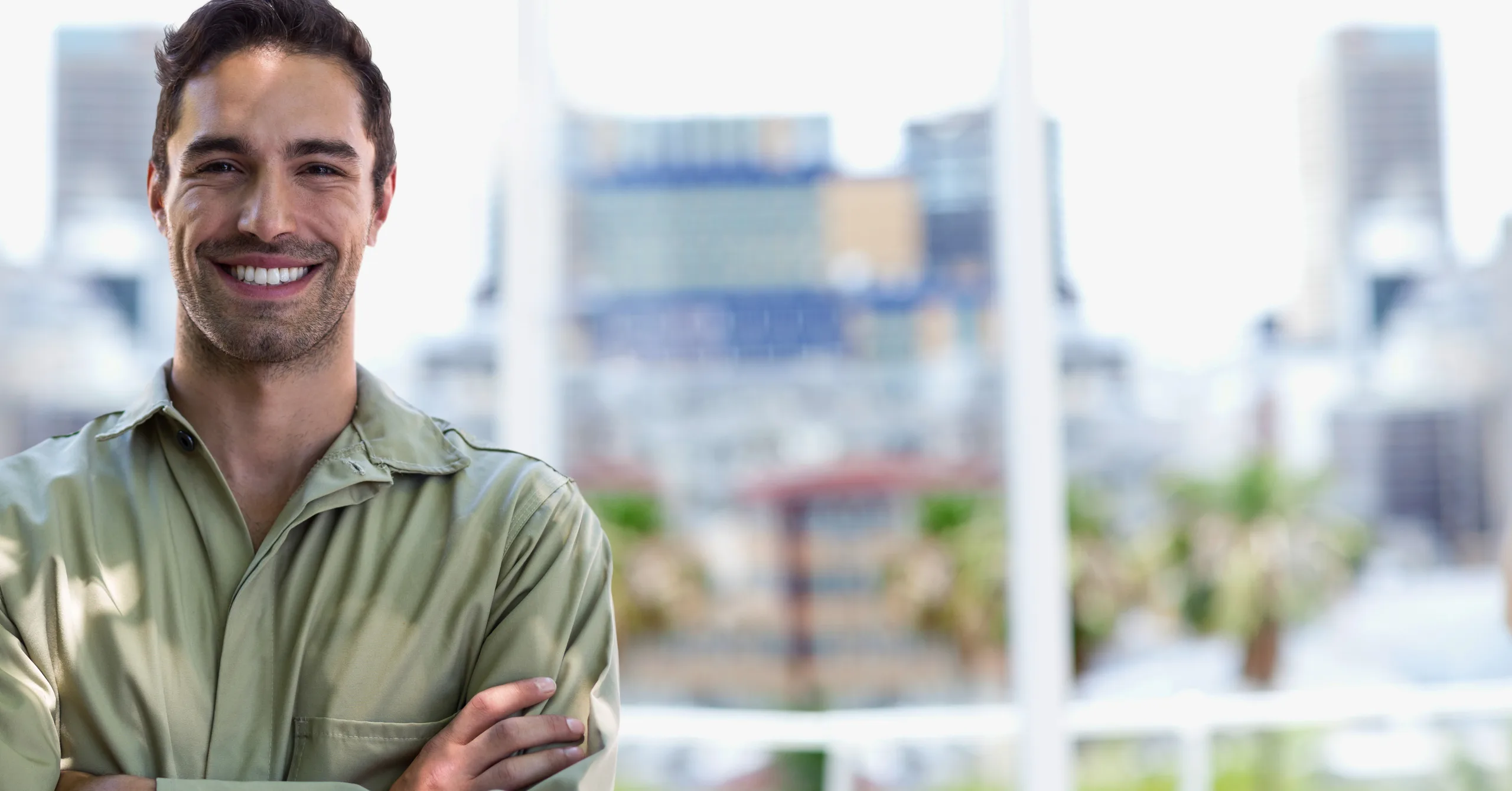 A young man in an olive green shirt smiles with his arms crossed in front of a large window; behind him, out of focus, a city skyline with buildings and palm trees.