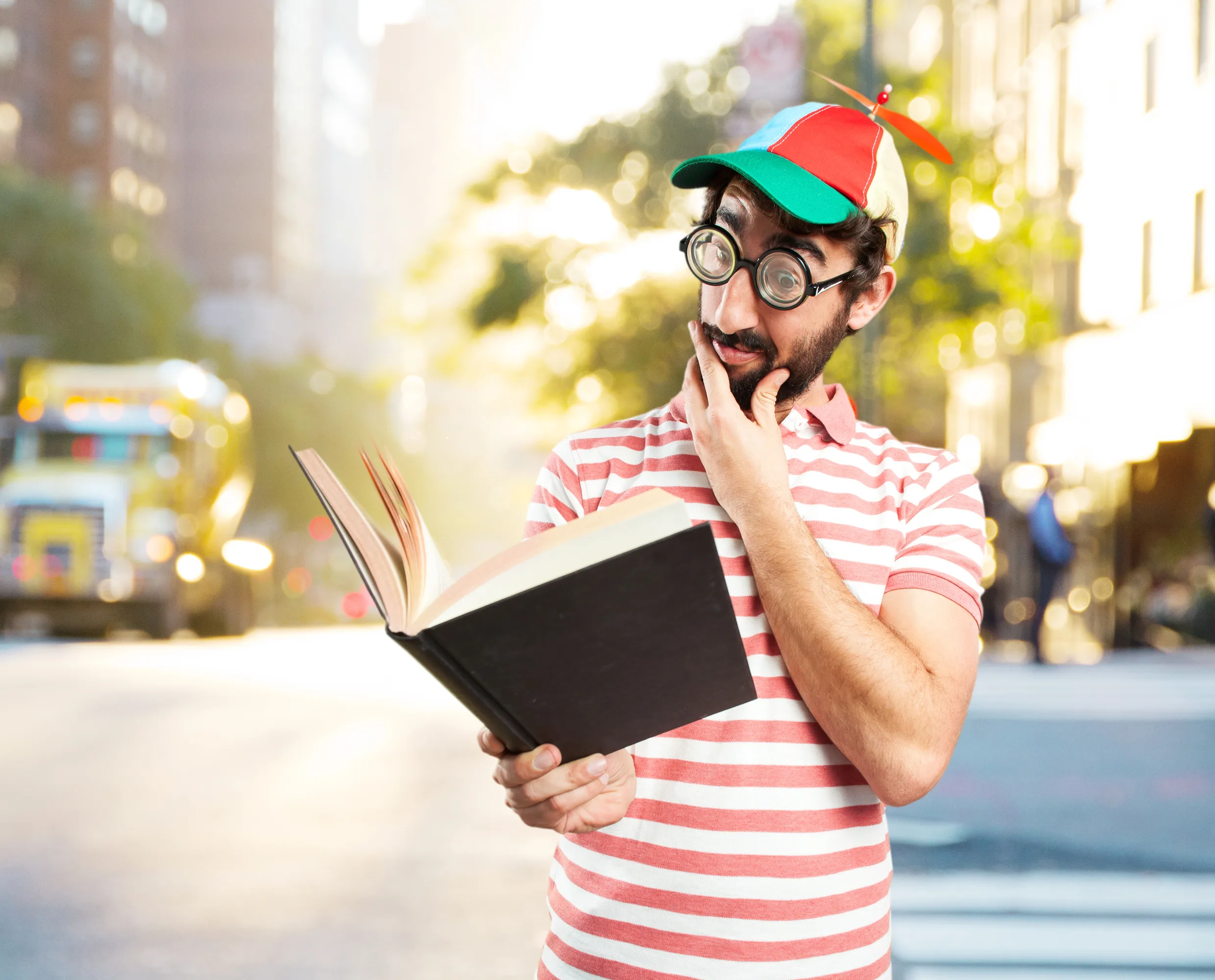 A young man in a colorful propeller cap and joke glasses holds an open book in the street with an amused, surprised expression.