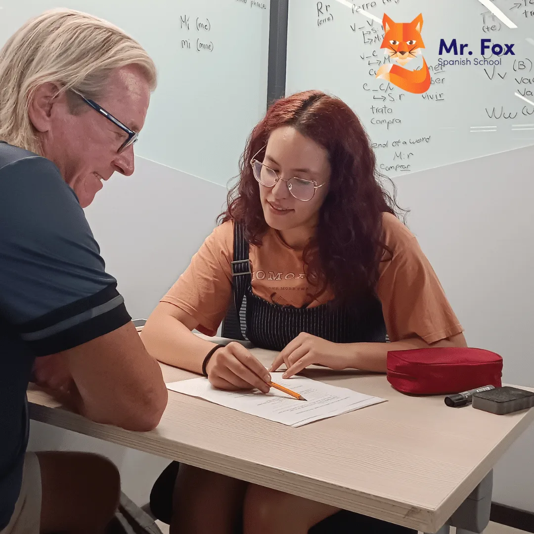 A young teacher goes over a worksheet with an older student seated at the same table, with a whiteboard behind them.