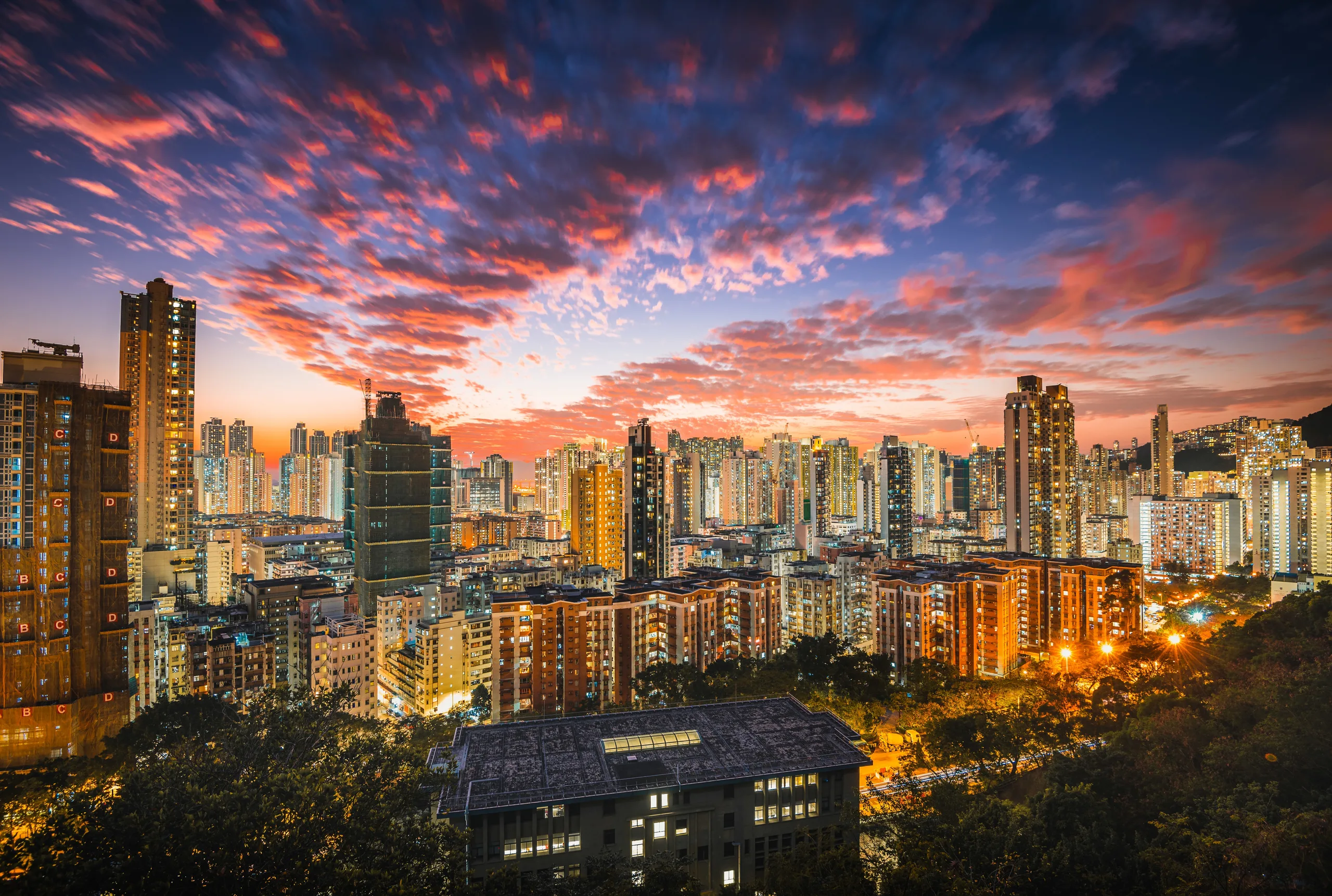 Panorámica nocturna de una gran ciudad asiática densamente poblada, con numerosos rascacielos y edificios residenciales iluminados bajo un espectacular cielo al atardecer con nubes